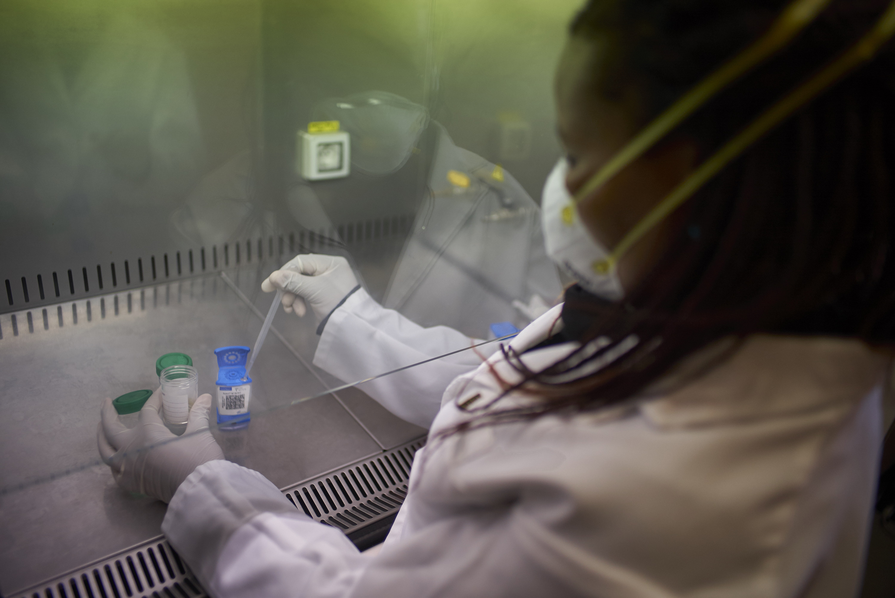 A researcher in a lab coat and mask dropping a solution into a container behind a protective window pane.